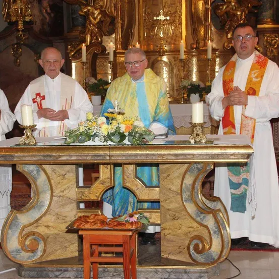 Drei Priester stehen an einem mit Blumen und Kerzen geschmückten Altar, einer hält ein Buch. Der Altar ist mit Gold und Marmor verziert.