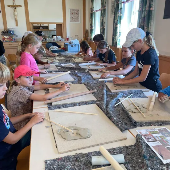 Eine Gruppe von Kindern sitzt an einem langen Tisch und ist mit einer Tonmodellierungsaktivität beschäftigt. Sie verwenden verschiedene Werkzeuge und Materialien auf ihren Tischen. Der Raum hat ein religiöses Symbol an der Wand.
