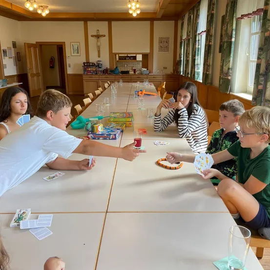 Eine Gruppe von Kindern und einer erwachsenen Frau sitzen um einen langen Tisch und spielen ein Kartenspiel. Der Raum hat Holzwände und große Fenster mit Vorhängen.