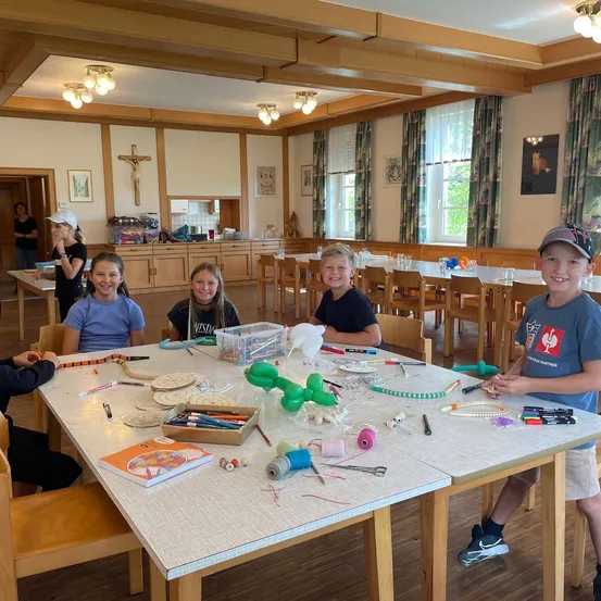 Fünf Kinder sitzen in einem Klassenzimmer um einen Tisch herum und beschäftigen sich mit einer Bastelaktivität. Verschiedene Bastelmaterialien liegen auf dem Tisch, darunter Luftballons, Perlen und Bücher. Ein Kreuz ist an der Wand hinter ihnen zu sehen.