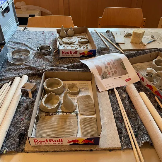 Ein Tisch mit verschiedenen Tonkunst-Utensilien, einschließlich einer Box Red Bull Energy Drink, Tonformen und Werkzeugen.