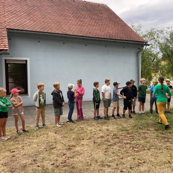 Eine Gruppe von Kindern steht in einer Reihe vor einem Gebäude mit einem roten Dach. Sie tragen verschiedenfarbige Hemden und Hosen.