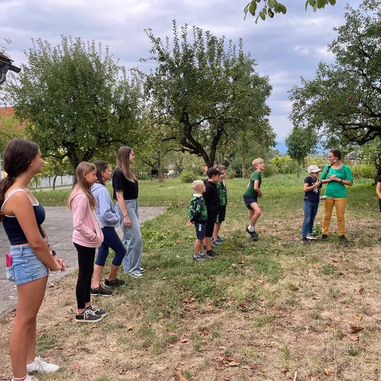 Eine Gruppe von Kindern und Erwachsenen steht auf einem Grasfeld mit Apfelbäumen im Hintergrund.