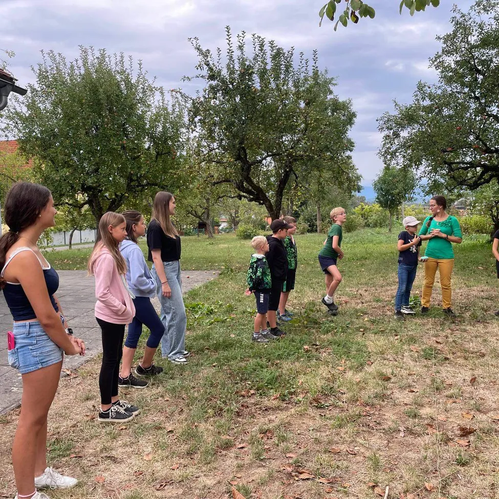 Eine Gruppe von Kindern und Erwachsenen steht auf einem Grasfeld mit Apfelbäumen im Hintergrund.