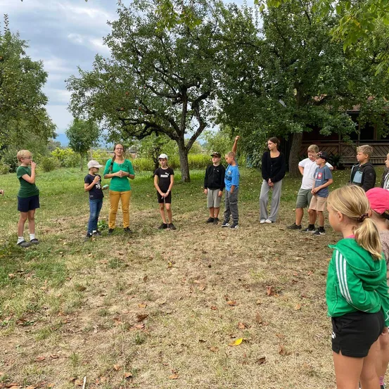 Eine Gruppe von Kindern und Erwachsenen steht auf einem Feld mit Bäumen im Hintergrund. Die Frau in Grün spricht zu der Gruppe.