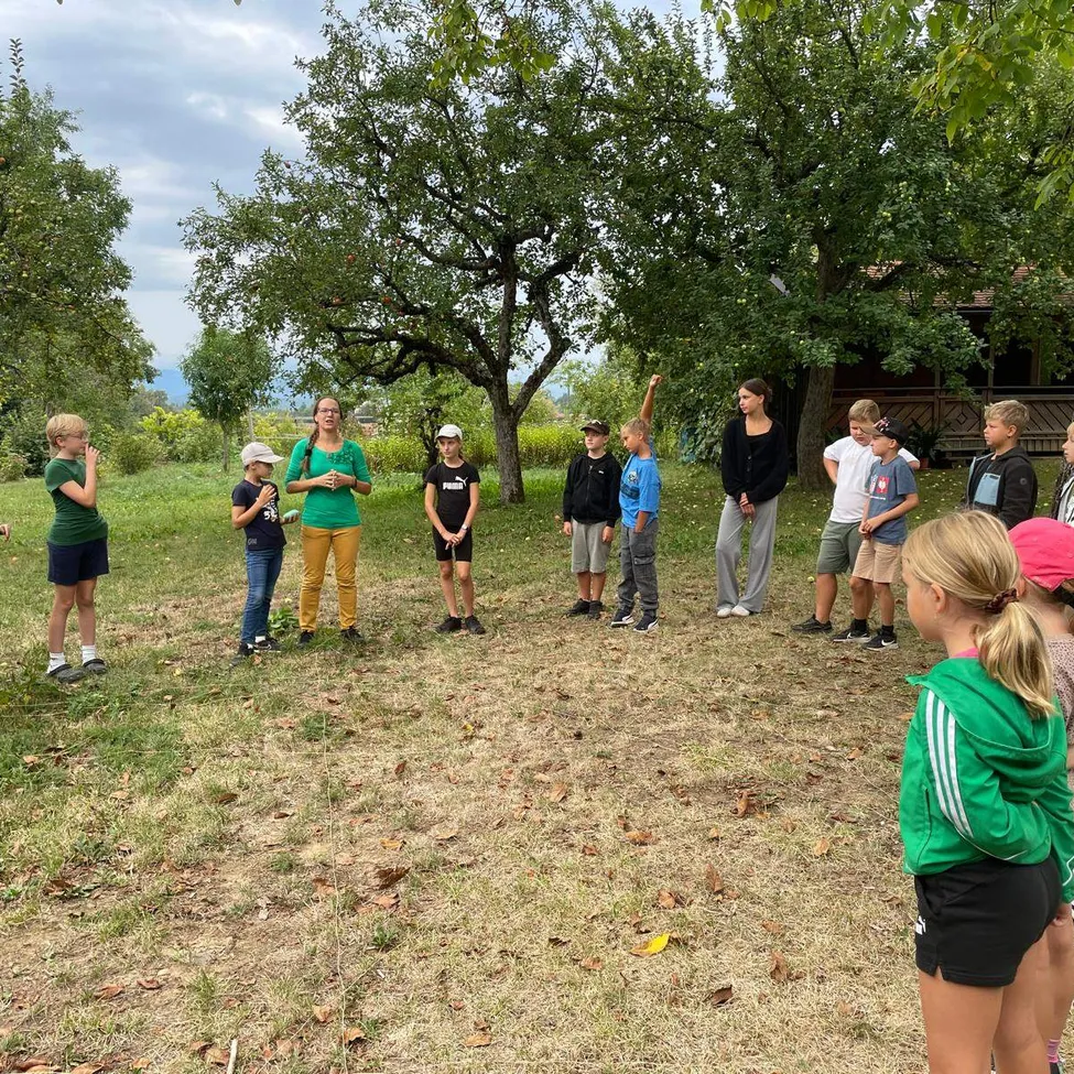 Eine Gruppe von Kindern und Erwachsenen steht auf einem Feld mit Bäumen im Hintergrund. Die Frau in Grün spricht zu der Gruppe.