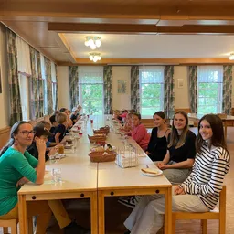 Eine Gruppe von Menschen sitzt an einem langen Tisch in einem Esszimmer, einige essen, während andere in die Kamera schauen. Es gibt Körbe mit Essen auf dem Tisch.