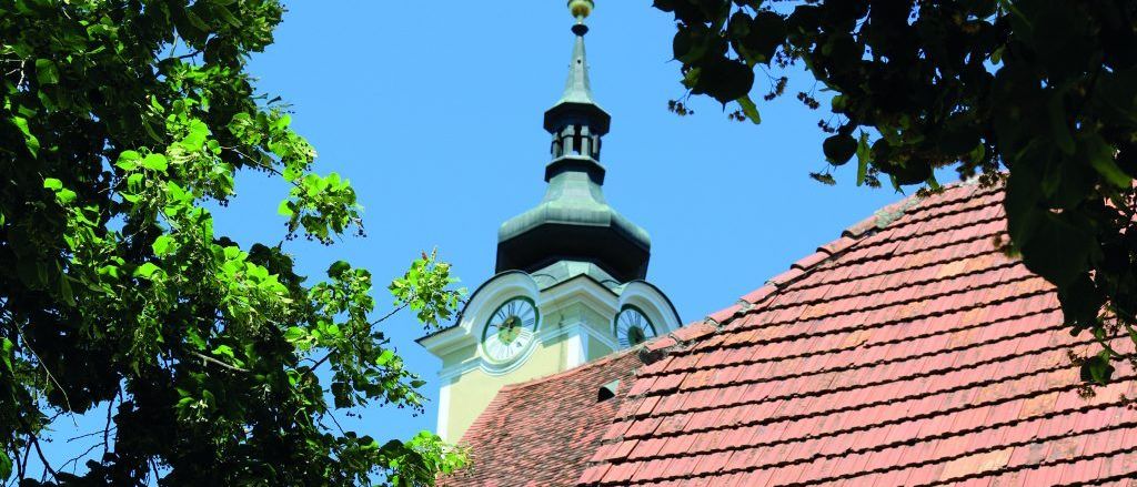 Ein Gebäude mit rotem Dach und einem Turm mit einer Uhr, umgeben von Bäumen an einem sonnigen Tag.