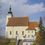 Pfarre Preding-Logo. Eine große Kirche mit braunem Dach und einem Kirchturm, umgeben von Bäumen und einem kleinen Ziegelweg. Die Kirche hat Rundbogenfenster und einen kleinen Eingang mit Treppen.