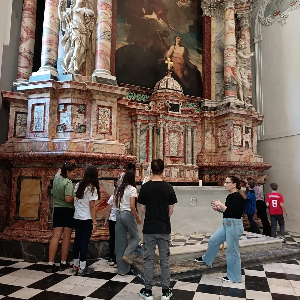 Eine Gruppe von Menschen steht vor einem Marmoraltar in einer Kirche. Der Altar hat ein Gemälde einer Frau, die ein Kreuz hält, und eine Statue eines Engels oben.