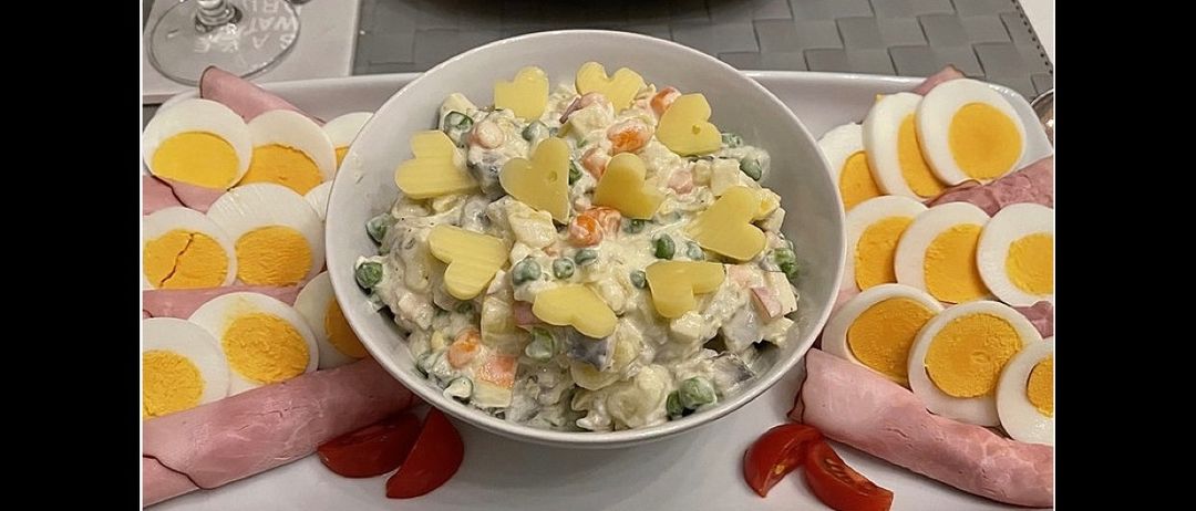 Ein weißer Teller hat eine Schüssel mit gelbem Salat in der Mitte, umgeben von geschnittenem Schinken und roten Tomaten. Der Salat ist herzförmig und mit gelben und weißen Zutaten garniert.