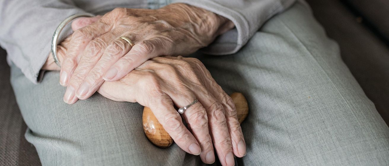 Eine ältere Person mit faltigen Händen hält einen hölzernen Stock in der rechten Hand und stützt die linke Hand darauf.