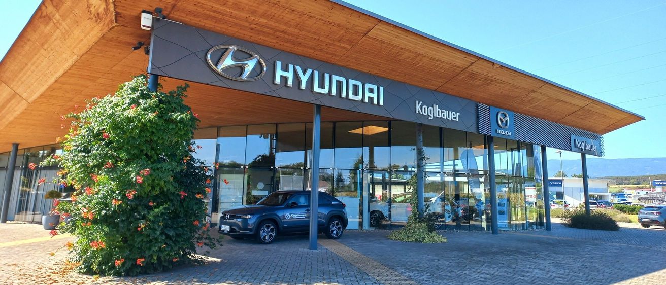 Außenansicht eines Hyundai-Autohauses mit einem dunkelblauen Hyundai SUV vor dem Gebäude. Das Gebäude hat ein modernes Holzdesign und Glasfenster.