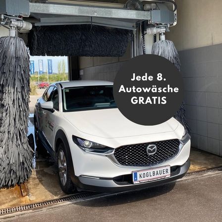 Ein weißer Mazda-Wagen wird in einer automatischen Waschanlage gereinigt. Die Waschanlage hat ein Schild mit der Aufschrift 'Jeder 8. Autowasche GRATIS'. Die Waschanlage ist mit Bürsten und einem Wassersprühsystem ausgestattet. Es gibt Banner an der Wand.