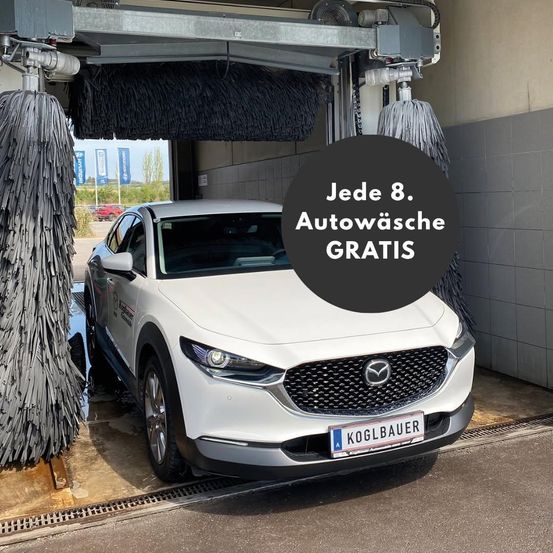 Ein weißer Mazda-Wagen wird in einer automatischen Waschanlage gereinigt. Die Waschanlage hat ein Schild mit der Aufschrift 'Jeder 8. Autowasche GRATIS'. Die Waschanlage ist mit Bürsten und einem Wassersprühsystem ausgestattet. Es gibt Banner an der Wand.