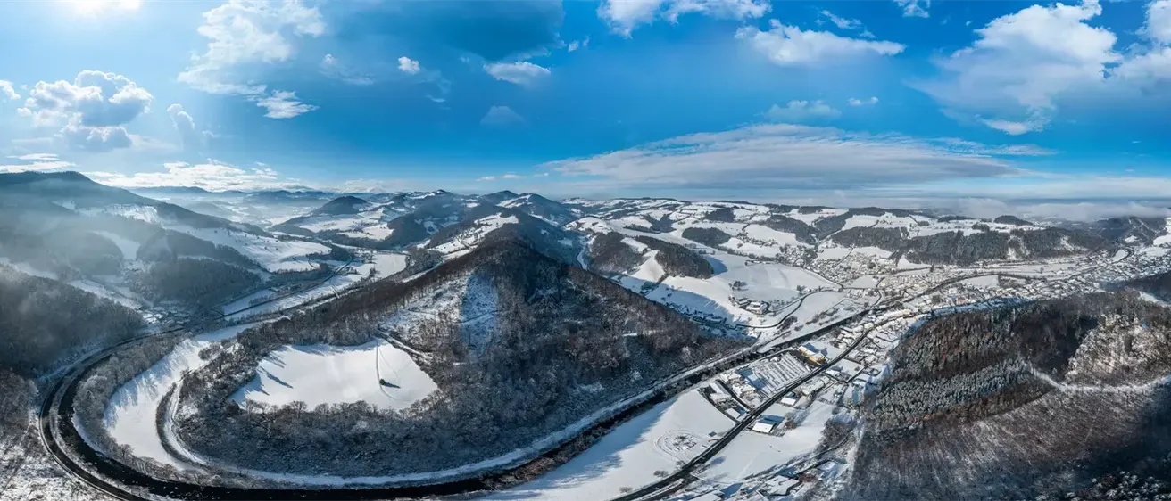 Luftaufnahme einer verschneiten Bergregion unter einem blauen Himmel mit verstreuten Wolken, die eine Stadt zwischen den verschneiten Hügeln und eine kurvenreiche Straße, die sich durch die Landschaft schlängelt, zeigt.