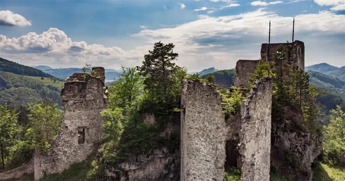 Auf einem felsigen Felsen stehen Überreste einer Burg mit klarem blauen Himmel und Wolken darüber. Bäume und Pflanzen wachsen durch die Ruinen.