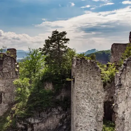 Auf einem felsigen Felsen stehen Überreste einer Burg mit klarem blauen Himmel und Wolken darüber. Bäume und Pflanzen wachsen durch die Ruinen.