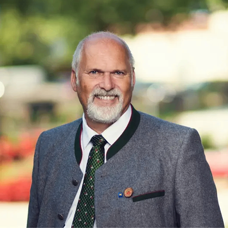 Ein Mann mit weißem Bart und Schnurrbart steht lächelnd in einem Park. Er trägt einen grauen Anzug mit einer grünen Krawatte und einer Anstecknadel auf der Brust.