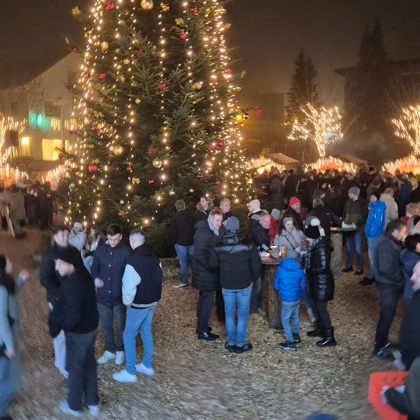 Bei Nacht ist ein großer Weihnachtsbaum mit bunten Lichtern und Ornamenten geschmückt. Eine Menschenmenge versammelt sich darum, einige stehen und andere sitzen an Tischen. Sie sind für das kalte Wetter warm gekleidet.