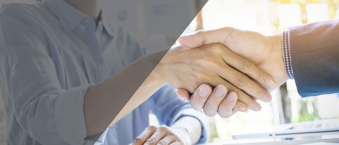 Zwei Personen in hellblauen Hemden schütteln sich die Hände, wahrscheinlich in einem professionellen Umfeld. Die Hände sind scharf, der Hintergrund verwischt.