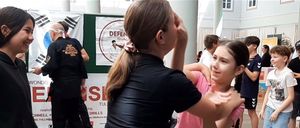 Ein junges Mädchen übt Kampfkunst mit einer weiblichen Ausbilderin. Hinter ihnen beschäftigen sich Menschen mit verschiedenen Aktivitäten, und ein Mann in Schwarz steht in der Nähe eines Schildes.