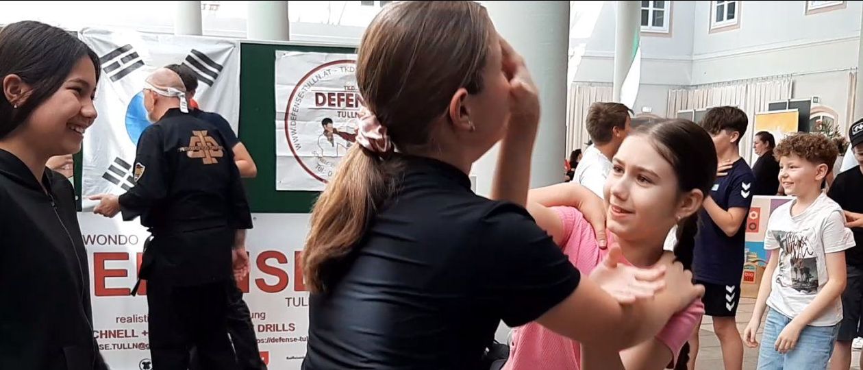 Ein junges Mädchen übt Kampfkunst mit einer weiblichen Ausbilderin. Hinter ihnen beschäftigen sich Menschen mit verschiedenen Aktivitäten, und ein Mann in Schwarz steht in der Nähe eines Schildes.