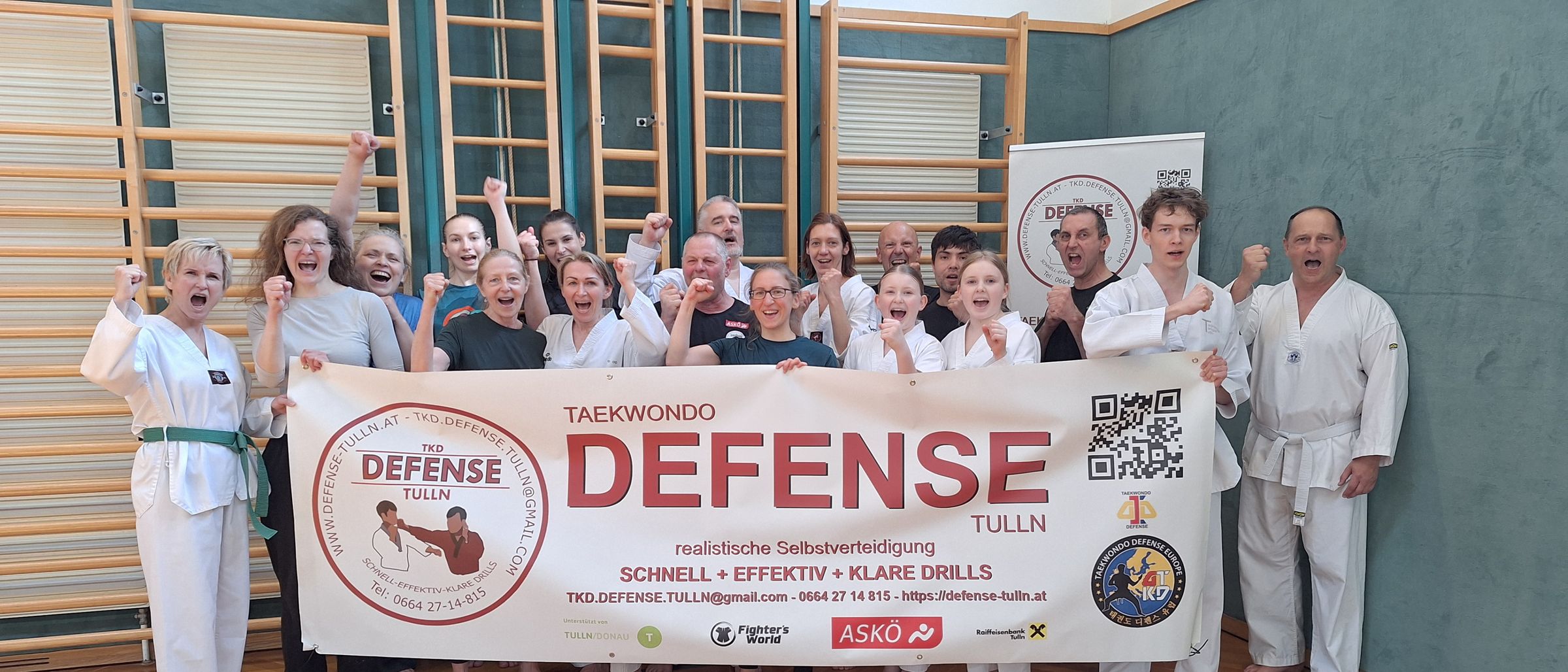 Eine Gruppe von Menschen in Kampfsportuniformen posiert mit einem Banner, auf dem 'Taekwondo Defense Tulln' steht. Sie stehen in einem Fitnessstudio mit Holzböden und einer hölzernen Wand im Hintergrund.