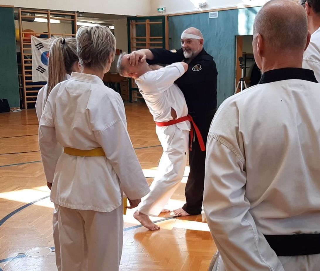 Ein Kampfsportlehrer demonstriert eine Technik an einem Schüler, während andere in einer Turnhalle zuschauen.