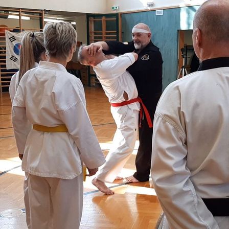 Ein Kampfsportlehrer demonstriert eine Technik an einem Schüler, während andere in einer Turnhalle zuschauen.