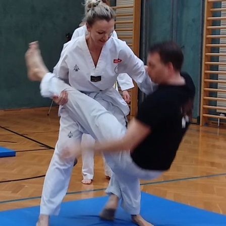 Eine Gruppe von Menschen übt Kampfsport in einer Turnhalle. Eine Frau führt einen Tritt aus, während eine andere Frau ihr Bein hält. Ein Mann liegt auf dem Boden, und ein Mann mit gelbem Gürtel beobachtet.