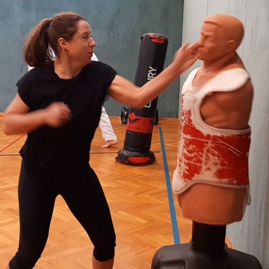 Eine Frau in einem Fitnessstudio übt Kampfsport, indem sie eine Puppe mit einem roten und weißen Westchen schlägt. Ein Boxsack ist im Hintergrund.