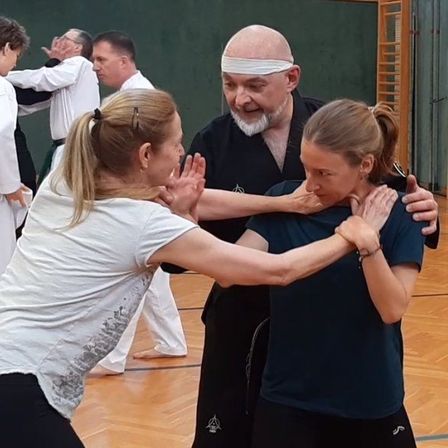 In einer Turnhalle unterrichtet ein Mann in schwarzer Kleidung zwei Frauen in weißen und blauen Shirts in Kampfkunsttechniken. Sie alle lächeln und scheinen die Lektion zu genießen.