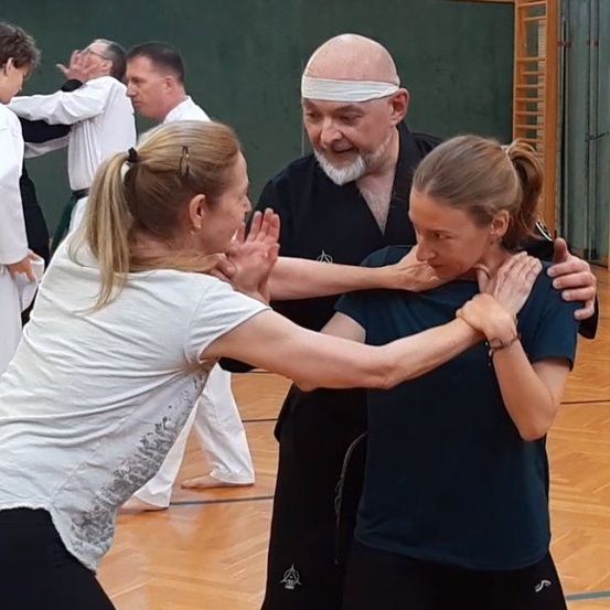 In einer Turnhalle unterrichtet ein Mann in schwarzer Kleidung zwei Frauen in weißen und blauen Shirts in Kampfkunsttechniken. Sie alle lächeln und scheinen die Lektion zu genießen.