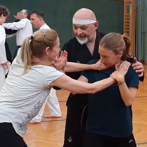 In einer Turnhalle unterrichtet ein Mann in schwarzer Kleidung zwei Frauen in weißen und blauen Shirts in Kampfkunsttechniken. Sie alle lächeln und scheinen die Lektion zu genießen.