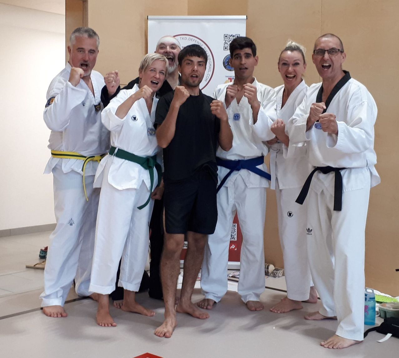Eine Gruppe von Menschen in Kampfsportuniformen posiert für ein Foto, alle tragen Gürtel außer dem zentralen Mann in Schwarz.
