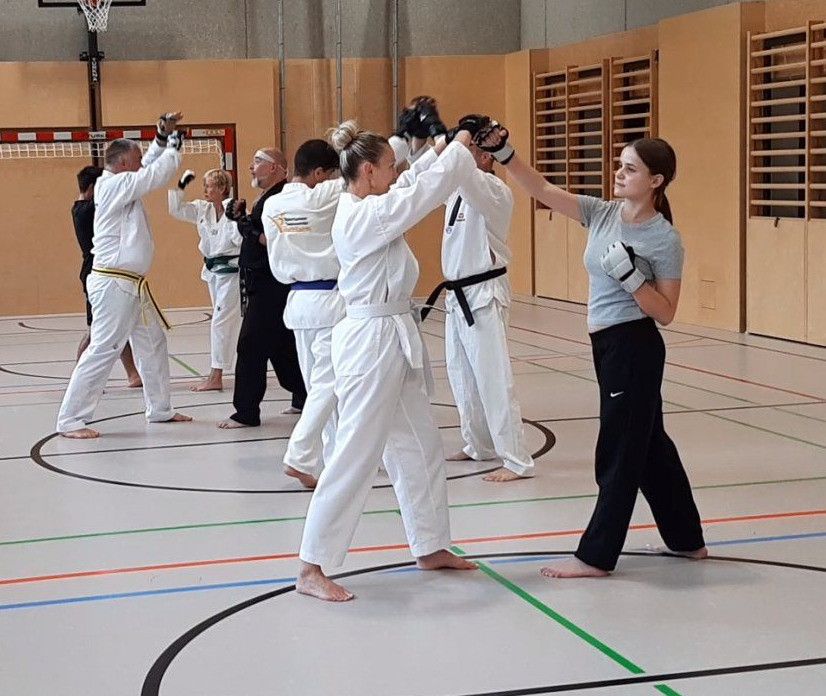 In einem Fitnessstudio üben eine Gruppe von Menschen in weißen Kampfsportuniformen Karate. Eine Frau in einem grauen Shirt und schwarzen Hosen leitet die Sitzung.