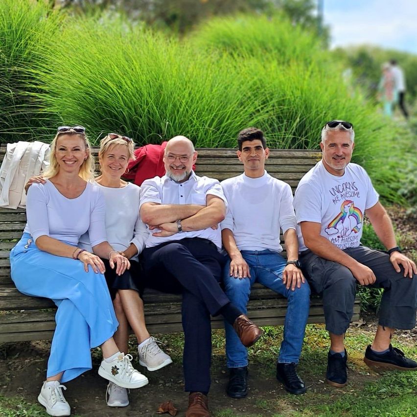 Fünf Menschen sitzen auf einer Holzbank in einem Park. Drei Männer und zwei Frauen. Sie alle lächeln. Der Mann auf der rechten Seite hat ein weißes T-Shirt mit einem Einhorn-Design. Der Hintergrund ist unscharf.