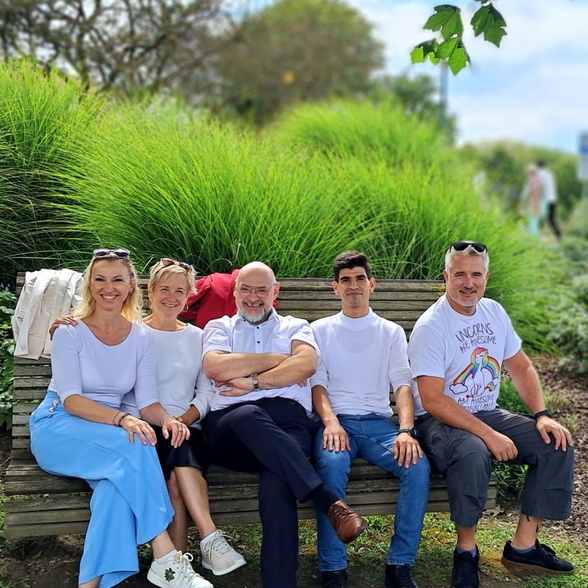 Fünf Personen sitzen auf einer Holzbank in einem Park. Zwei Frauen und drei Männer, einer trägt ein T-Shirt mit dem Wort 'Einhörner' darauf.