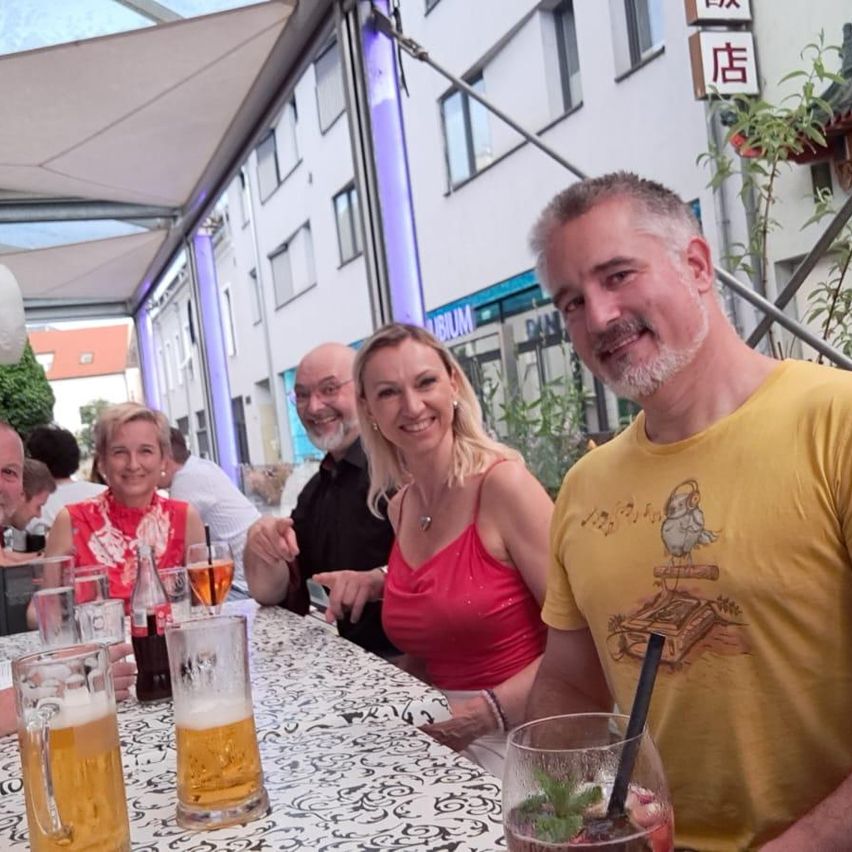 Eine Gruppe von Menschen sitzt an einem Bartisch, lächelt und posiert für ein Foto. Sie haben Biergläser, Flaschen und ein Getränk mit einem Strohhalm. Der Mann trägt ein gelbes T-Shirt mit grafischem Design. Hinter ihnen steht ein Gebäude mit chinesischen Schriftzeichen an der Wand.