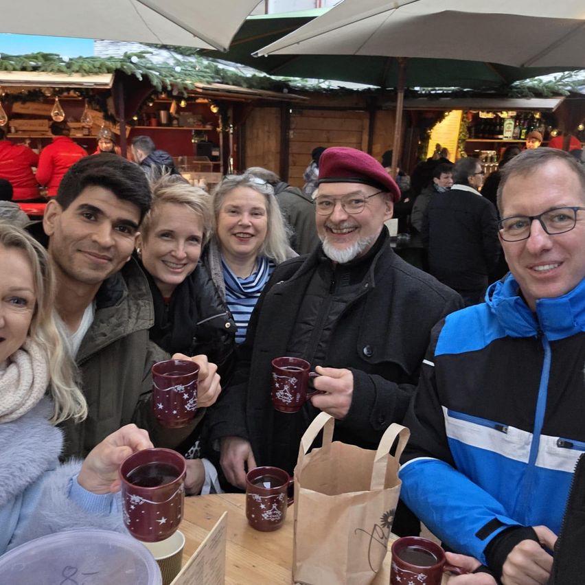 Fünf Personen sitzen an einem Tisch mit Kaffeetassen und lächeln für ein Foto auf einem festlichen Markt. Dahinter sind Stände mit Schirmen für Weihnachten dekoriert.