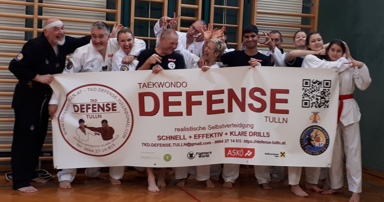 Eine Gruppe von Personen in weißen Uniformen posiert mit einem großen Banner, auf dem 'Taekwondo Defense Tulln' steht. Sie lächeln und scheinen an einer Kampfsportveranstaltung teilzunehmen. Das Banner enthält Kontaktinformationen und eine Website.