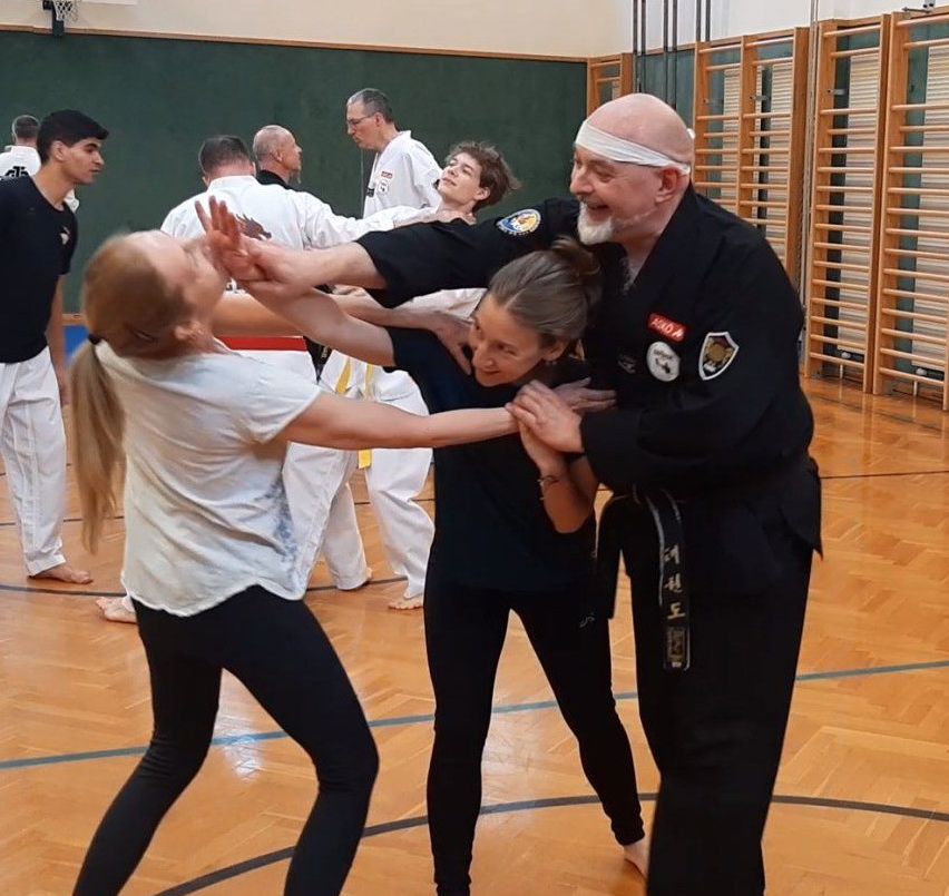In einer Turnhalle unterrichtet ein Mann zwei Frauen im Karate. Eine Frau trägt ein weißes Shirt, die andere ein schwarzes. Dahinter üben andere Personen Karate.