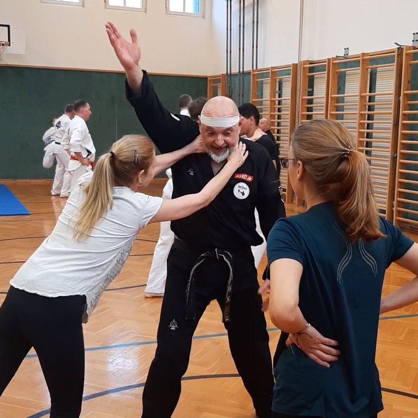 Ein älterer Mann in einem schwarzen Kampfsportanzug trainiert mit zwei Frauen in einer Turnhalle, andere im Hintergrund.