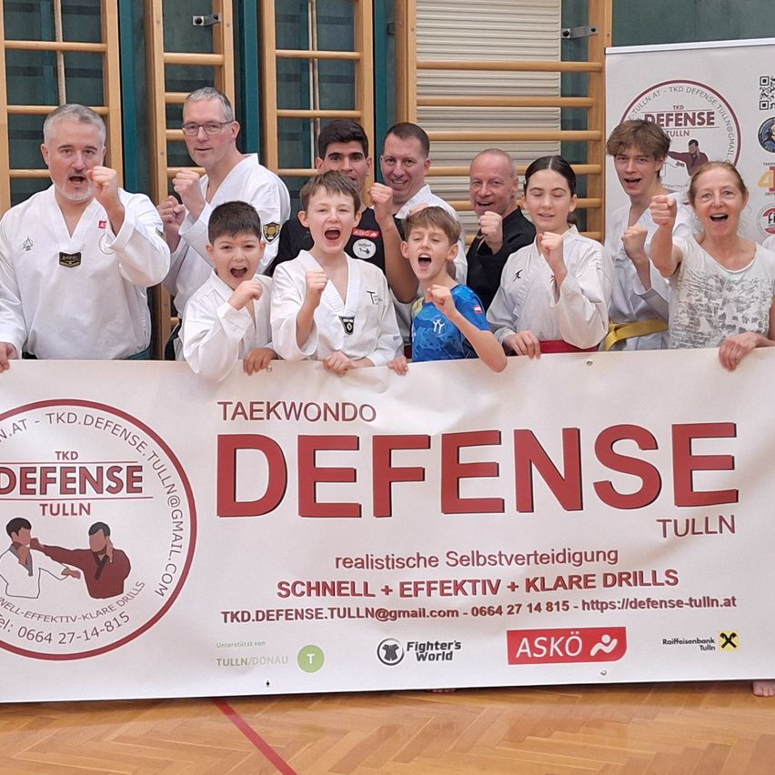 Eine Gruppe von Menschen in weißen Kampfsportuniformen posiert für ein Foto vor einem großen Banner. Das Banner steht in rot und enthält Informationen über die TKD Defense Tulln Schule.