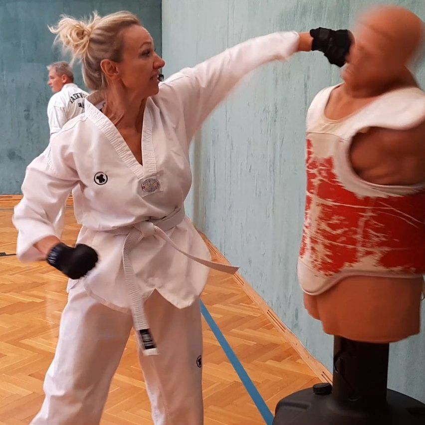 Eine Frau in einem weißen Karate-Anzug übt ihre Schläge an einem Mannequin, das eine rote Weste trägt. Sie trägt Handschuhe. Hinter ihr steht eine weitere Person. Der Raum hat Holzböden und eine hellblaue Wand.
