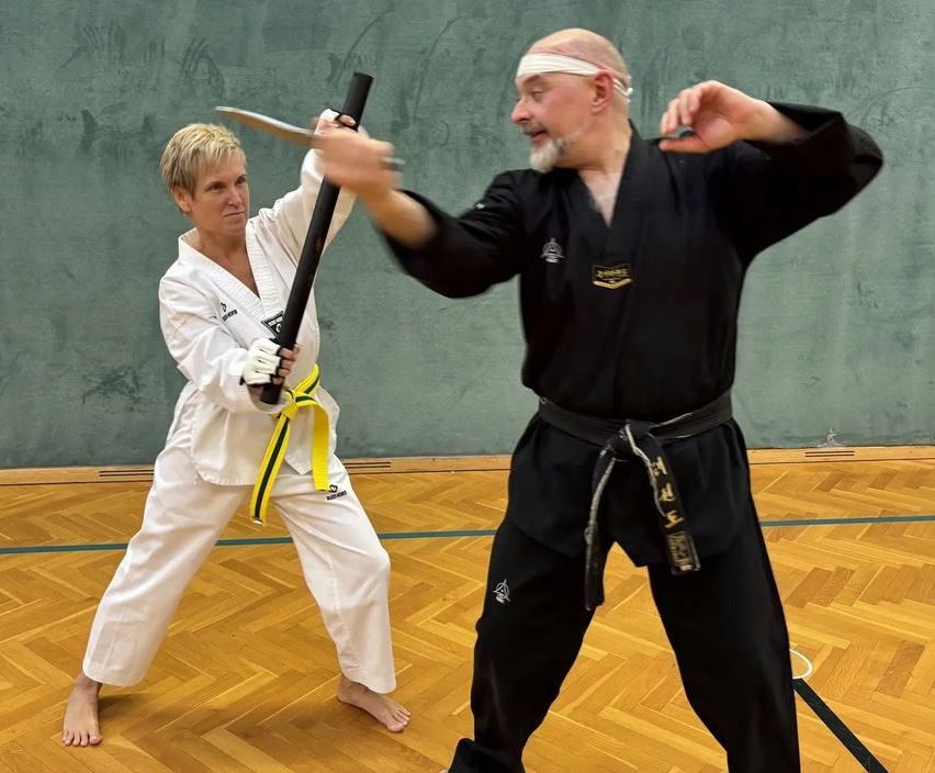 Zwei Personen in Kampfsportuniformen üben Kampftechniken in einem Fitnessstudio. Eine Frau in Weiß hält ein Schwert, während ein Mann in Schwarz einen Stock hat.