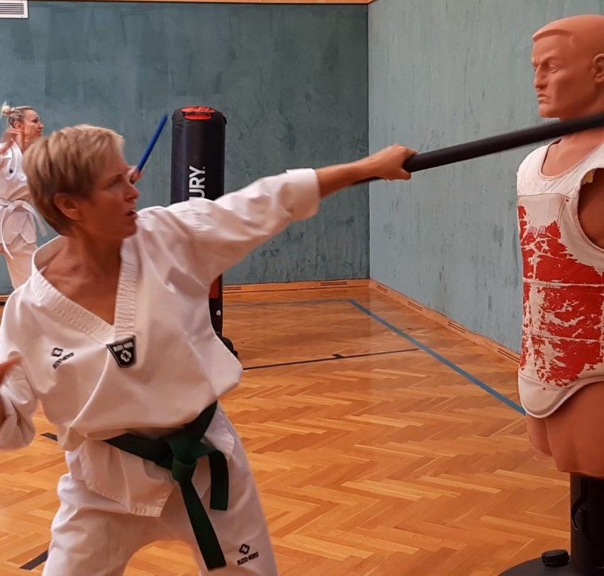 An older woman in a white karate uniform practices her strikes on a mannequin wearing a red and white vest in a gym.
