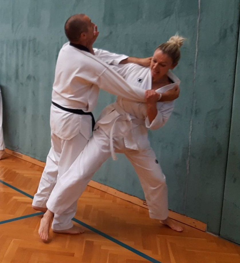 Two individuals, dressed in white martial arts uniforms, engage in a martial arts technique. The person on the left has a black belt and is holding the person on the right. They are in a fighting stance, possibly practicing judo.