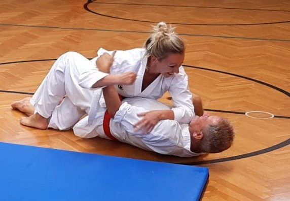 A woman in a white martial arts uniform is on top of a man practicing judo on a wooden floor.